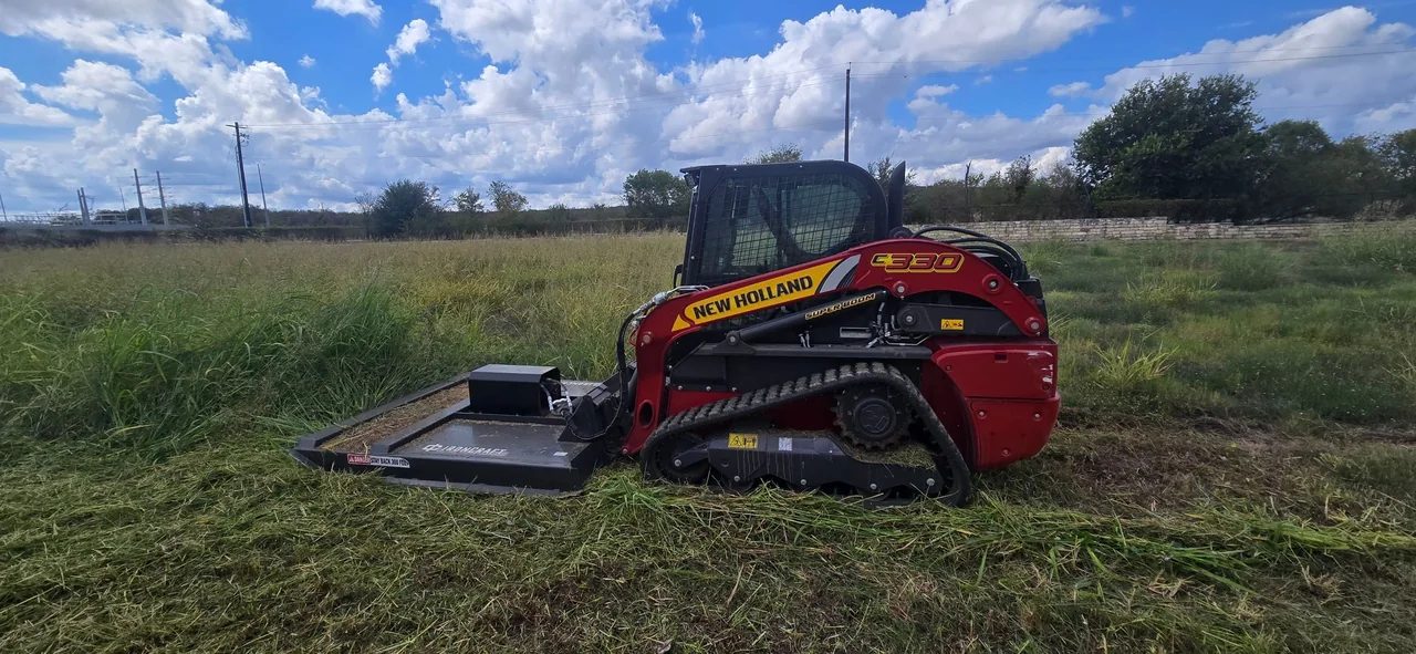 Tracked loader fitted with a brush-cutting attachment in an open grassy area.
